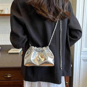 Puede incluir: Bolso plateado metálico con cordón y correa de cadena. El bolso tiene una parte superior fruncida y un pequeño emblema decorativo en la parte delantera. La correa de cadena es plateada y parece estar hecha de pequeñas piezas de metal unidas.