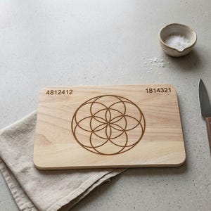 Könnte beinhalten: Ein helles Holzschneidebrett mit einem geometrischen Design und den eingravierten Zahlen "4812412" und "1814321". Eine kleine Keramikschale mit Salz, ein Messer und eine Leinen-Serviette sind ebenfalls vorhanden.