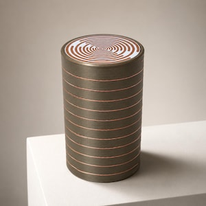 Könnte beinhalten: Ein zylindrischer Behälter mit dunkler Metalloberfläche und horizontalen kupferfarbenen Ringen. Der Deckel hat eine weiße kreisförmige Oberfläche, die mit einem konzentrischen kupferfarbenen Spiralmuster verziert ist.