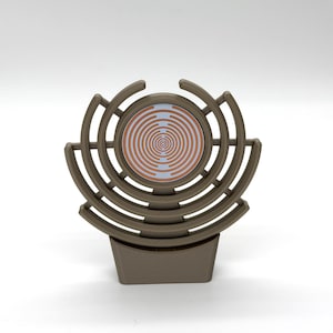 Op de afbeelding: Een bruin decoratief object met een cirkelvormig ontwerp. Het object heeft een vierkante basis en een reeks gebogen lijnen die een cirkelvormig patroon vormen rond een centrale schijf met een spiraalvormig ontwerp in koper- en wittinten.