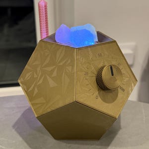 May include: A gold-colored, dodecahedron-shaped device with a textured geometric surface. It features a circular dial marked with numbers 0, 25, 50, 75, and 100. The top opening displays glowing blue and purple light, and a pink, textured rod extends upward.