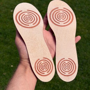 Op de afbeelding: Een paar lichtbeige leren schoenzolen met twee in reliëf aangebrachte koperkleurige cirkelvormige doolhofpatronen op elk stuk, één bij de hiel en één bij de voorvoet, met een gladde, natuurlijke textuur en afgeronde randen.