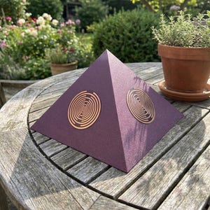 Op de afbeelding: Een donkerpaars piramidevormig object met een textuur op het oppervlak. Twee van de zichtbare driehoekige zijden zijn voorzien van een gecentreerde, koperkleurige cirkelvormige afbeelding bestaande uit meerdere concentrische ringen die naar het midden spiraliseren.