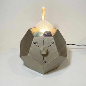 Puede incluir: Un dispositivo gris de forma geométrica con un dial marcado 10, 50 y 100. El dispositivo tiene una luz en la parte superior, con un cristal transparente y dos piedras de colores en el interior. Una pieza de metal enrollada se extiende desde la parte superior.