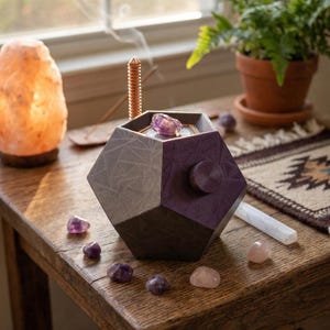May include: A geometric, two-toned aromatherapy diffuser with a purple top and a gray base, topped with a purple crystal. Surrounding the diffuser are small amethyst and rose quartz crystals, and a lit Himalayan salt lamp.