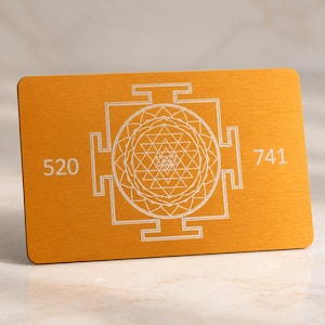 Peut inclure: Une carte rectangulaire de couleur dorée présentant un motif géométrique Sri Yantra gravé en blanc au centre. Le nombre 520 est imprimé sur le côté gauche du motif, et le nombre 741 est imprimé sur le côté droit du motif.