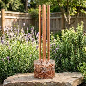 Peut inclure: Un carillon de guérison sonore de couleur cuivre avec quatre tubes verticaux s'élevant d'une base transparente remplie de cristaux et de copeaux de cuivre. Le carillon est posé sur une pierre dans un jardin avec de la lavande et de la verdure.