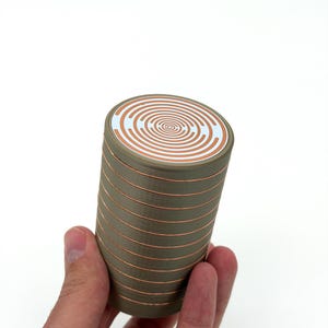 Könnte beinhalten: Zylindrisches Objekt mit einem Spiralmuster auf der Oberseite. Das Objekt ist olivgrün mit kupferfarbenen Bändern. Die Oberseite hat ein weißes und kupferfarbenes Spiralmuster. Das Objekt wird in einer Hand vor weißem Hintergrund gehalten.