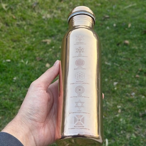 May include: A metallic copper-colored water bottle featuring a series of six stacked geometric symbols and numerical codes printed in black. Each symbol is accompanied by text labels including Spiritual Connection, Higher Intuition, and Protection.