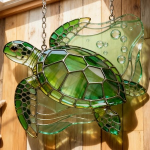 Puede incluir: Un atrapasoles de vidriera de tortuga marina con tonos verdes y vidrio transparente. La tortuga está suspendida por una cadena, con un fondo de vidrio transparente y burbujeante. El atrapasoles cuelga frente a una pared de madera.