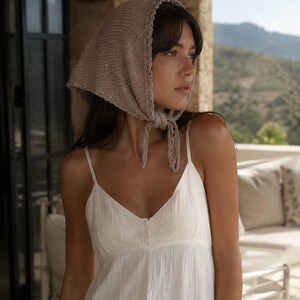 May include: A beige knitted headscarf with a delicate scalloped edge and tie closure, paired with a white sleeveless dress featuring a V-neckline and a button-down front detail. The headscarf is textured with a fine knit pattern and sits securely.
