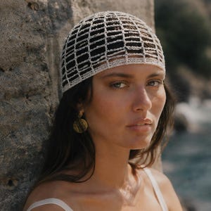 May include: A white crocheted head covering with a grid-like pattern. The head covering is worn with gold-colored circular earrings. The background features a stone wall and a blurred view of the ocean.