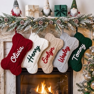May include: Five bone-shaped knit Christmas stockings hanging in a row. The stockings are red, white, beige, gray, and dark green, each featuring a cable knit texture and a personalized name embroidered in a contrasting color: Olivia, Henry, Evelyn, Wesley, and Tommy.