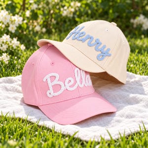 Op de afbeelding: Twee baseballpetten, een lichtroze met witte touw-stijl letters Bella en een crèmekleurige met lichtblauwe touw-stijl letters Henry. Beide petten hebben een gestructureerde kroon, een gebogen klep en kleine ventilatiegaatjes.