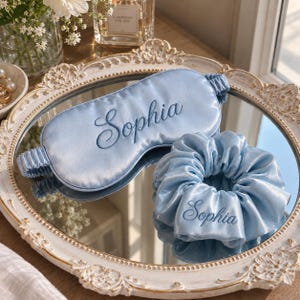 May include: A light blue satin sleep mask and a matching light blue satin scrunchie. Both items feature the name Sophia embroidered in a dark blue cursive script. The sleep mask has an elasticated strap, and the scrunchie is gathered fabric.