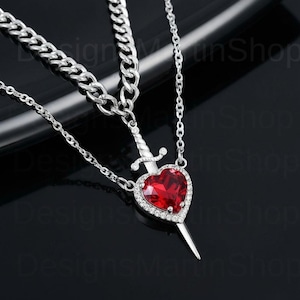 May include: A silver-toned layered necklace featuring a heart-shaped pendant pierced by a sword. The heart is a vibrant red gemstone, surrounded by a halo of small, clear stones. The necklace has a chain and a thicker curb chain.