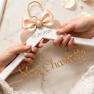 Peut inclure: Un cintre de robe de mariée en bois blanc avec un crochet doré et un nœud en satin. Le nœud est orné d'un bijou en forme de perle. Le cintre est personnalisé avec la date 05.20.2026 et le nom "Mrs. Charlotte" en fil d'or.