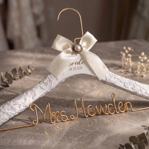 Peut inclure: Un cintre en bois blanc recouvert de dentelle, doté d'un crochet en métal doré et d'un nœud en satin crème avec une perle centrale. Le cintre porte l'inscription "Bride" et "08.28.2026" en noir, avec "Mrs. Howden" formé en fil doré.