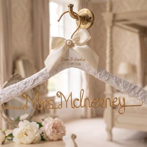 Peut inclure: Un cintre en bois blanc avec un motif floral texturé, doté d'un crochet en métal doré et d'une inscription en fil doré Mrs. McInerney. Un nœud en ruban de satin ivoire avec une perle centrale est fixé sous le crochet.