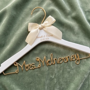 Peut inclure: Un cintre en bois blanc avec un crochet en métal doré et un nœud en ruban de satin crème. Le cintre présente le texte gravé Sarah & Davia 12/09/2026 et une inscription en fil métallique doré Mrs. McInerney sur la barre inférieure.
