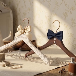 Peut inclure: Deux cintres en bois avec des crochets dorés. L'un est blanc avec une texture en dentelle, un nœud champagne et la date 05.20.2026. L'autre est en bois foncé avec un nœud bleu marine et deux boutons. Les noms Mrs. Charlotte et Mr. William sont en fil.