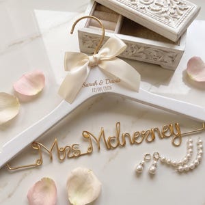 Peut inclure: Un cintre en bois blanc avec une inscription en fil métallique doré formant Mrs. McInerney, un ruban en satin crème noué au crochet et le texte imprimé Sarah & Davia 12/09/2026. Le cintre est accompagné d'une paire de boucles d'oreilles en perles.