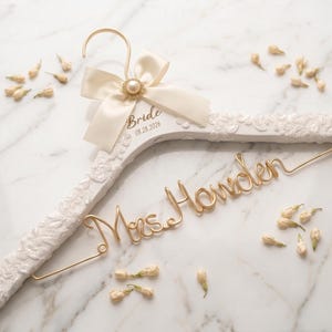 Puede incluir: Una percha de madera blanca con un revestimiento de encaje texturizado, que presenta una inscripción en alambre dorado que dice Mrs. Howden. La percha está adornada con un lazo de satén crema y una perla, con el texto Bride y 08.28.2026 impreso.