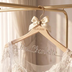 May include: Wooden wedding dress hanger with a cream-colored bow and a decorative jewel. The hanger has the date 05.20.2026 engraved on it, and the name "Mrs. Charlotte" is written in wire. A white lace dress hangs from the hanger.