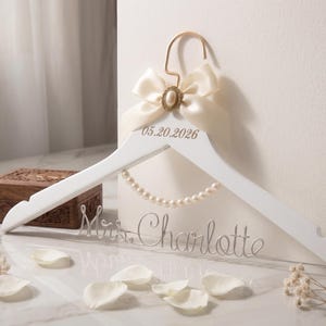 May include: White wooden wedding dress hanger with a cream-coloured bow and pearl accents. The hanger is personalised with the date 05.20.2026 and the name "Mrs. Charlotte" in cursive script. A decorative wooden box is in the background.