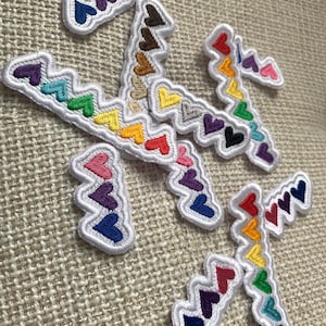 May include: Embroidered patches in various shapes, each featuring a row of colorful hearts. The hearts are in a rainbow of colors, including red, orange, yellow, green, blue, and purple, with white stitching.