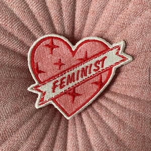 Peut inclure: Écusson rouge en forme de cœur avec le mot « FEMINIST » sur une bannière blanche. Le patch a un contour rouge et quatre éclats d'étoiles rouges. Le fond est un tissu rose texturé.