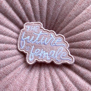 May include: A pink embroidered patch with the words "THE FUTURE IS FEMALE" in light blue script. The patch has a white border and is set against a textured pink fabric background.