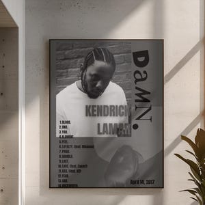 May include: Black and white framed poster featuring Kendrick Lamar. The poster includes the album title "DAMN." and tracklist. The album's release date, April 14, 2017, is also displayed. The image is displayed above a white sofa.
