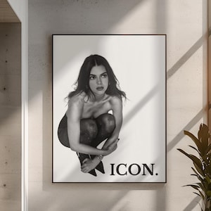 May include: Black and white poster featuring a person in a squatting pose. The person is wearing patterned tights and heels. The word "ICON." is printed in a bold font at the bottom of the poster. The poster is hanging on a wall above a white sofa.