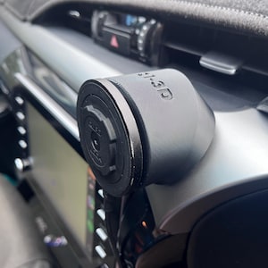 Puede incluir: Soporte negro para teléfono de coche con el texto "1-3D" impreso en el lateral. El soporte está fijado al salpicadero de un coche, con un enfoque en la base circular del soporte y el interior circundante.