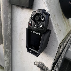 Puede incluir: Un mando a distancia negro de la marca Lowrance montado en un soporte de plástico negro. El dispositivo cuenta con un joystick central, un botón de más e iconos de una hélice y un ancla. El nombre Lowrance está impreso en blanco en el frente.
