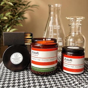 May include: Two amber glass candle jars with white labels reading Zoloft (Sertraline) and listing Bergamot, Sage, Rose Water, and Sandalwood. One jar is 8 oz (226 g). A black matchbox and a black lid with an Apothecary General logo are included.