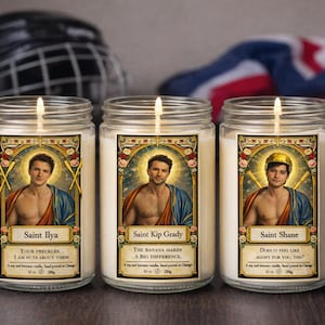 May include: Three glass jar candles with labels featuring illustrations of men in saintly attire. The labels read "Saint Ilya," "Saint Kip Grady," and "Saint Shane." Each candle has a lit wick and is surrounded by a hockey helmet and a flag.
