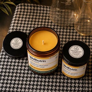 May include: Two amber glass jars containing yellow wax candles with black lids. The labels read Wellbutrin, Grapefruit, Bergamot, Ginger, Dry Cedar, 8 oz (226 g) Soy & Beeswax Candle. One lid is off, showing the candle wick and wax surface.