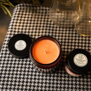 May include: An amber glass jar candle with orange wax and a single cotton wick, accompanied by two black lids featuring white circular labels that read Apothecary General Goods Store Chicago. The items are arranged on a black and white houndstooth fabric.
