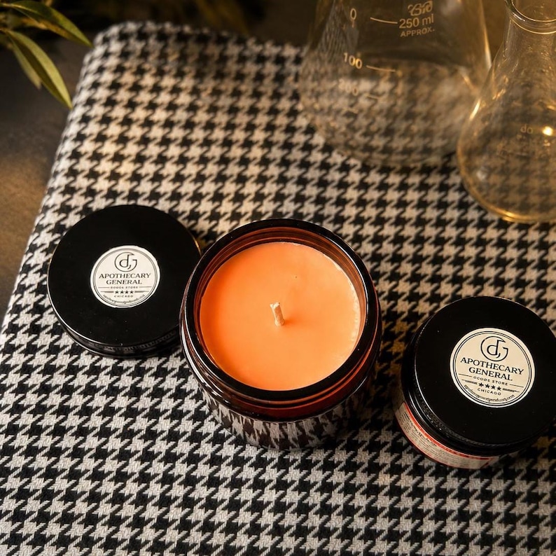 May include: An amber glass jar candle with orange wax and a single cotton wick, accompanied by two black lids featuring white circular labels that read Apothecary General Goods Store Chicago. The items are arranged on a black and white houndstooth fabric.