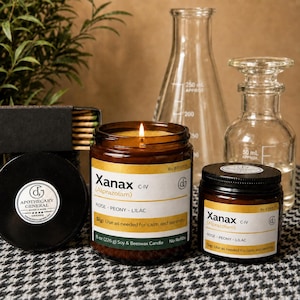 May include: Two amber glass candles with white labels reading Xanax (Alprazolam) Rose-Peony-Lilac, one lit and one lidded. Beside them are a black matchbox, a clear glass Erlenmeyer flask, and a clear glass apothecary bottle on a houndstooth surface.