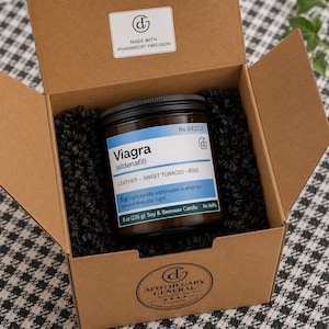 May include: A brown cardboard box containing a glass candle jar with a black lid. The label reads Viagra (sildenafil), Leather-Sweet Tobacco-Rose, 8 oz (226 g) Soy & Beeswax Candle. The box interior has black paper filler and a logo sticker.