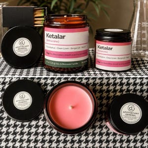 May include: Two amber glass jars containing pink soy and beeswax candles with labels reading Ketalar (Ketamine), Eucalyptus, Clean Linen, Bergamot, Musk, and Apothecary General. The jars have black lids and are shown from side and top views.