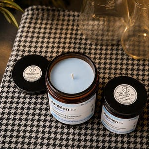May include: Two amber glass jars with black lids and light blue wax candles inside. The labels feature the text Ambien, Zolpidem, C-IV, Rx 591010, Chamomile, Moonflower, Sandalwood, Musk, and the Apothecary General logo with five stars.