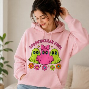 Può includere: Una felpa con cappuccio rosa chiaro con una stampa grafica di tre fantasmi e fiori. Il fantasma centrale è rosa con un fiocco, affiancato da due fantasmi verde lime. Il testo "SPOOKTACULAR VIBES" è stampato in rosa sopra i fantasmi.
