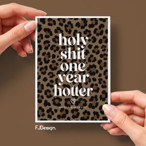 Könnte beinhalten: Eine rechteckige Grußkarte mit einem braun-schwarzen Leopardenmuster. Die Karte zeigt weißen Text mit der Aufschrift "holy shit one year hotter", darunter ein kleines Herzsymbol und die Worte "HAPPY BIRTHDAY" in kleineren weißen Buchstaben.