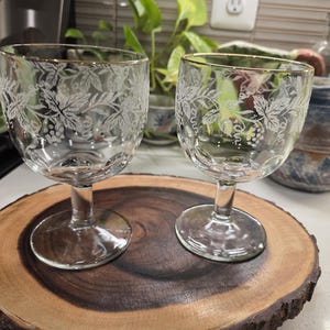 May include: Two clear wine glasses with gold rims and etched grape and leaf designs sit on a round, wooden serving board. The glasses have a short stem and a wide bowl. The board has a natural, rustic edge.
