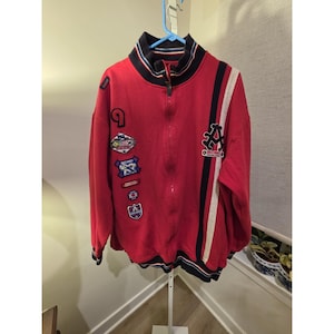Puede incluir: Una chaqueta roja con cremallera y cuello alto con ribete de rayas negras y blancas. La parte delantera muestra varios parches bordados, incluida una letra A grande, y rayas verticales negras y blancas en el lado izquierdo del pecho.