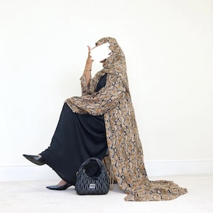May include: A person wearing a black dress and a long, snake-print patterned coat with a matching head covering. A black handbag with the brand name is on the floor. The person is wearing black shoes.